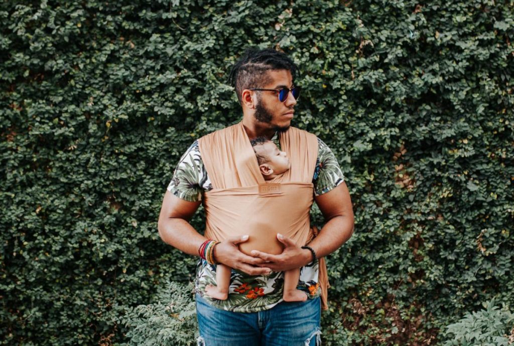 A man in a floral shirt wears a baby in a tan wrap to illustrate hot weather babywearing safety