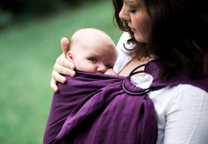 ASTM Baby Sling Standard is represented by a parent in a white shirt carrying a baby in a plum colored Bibetts sling